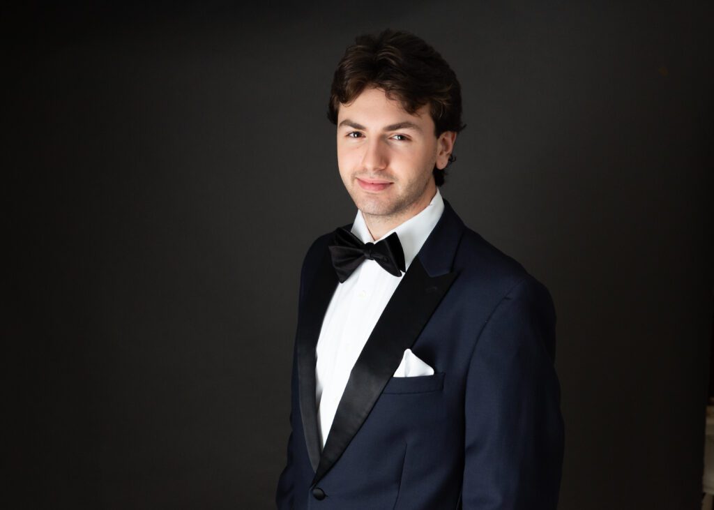 Studio portrait of a senior guy in a tuxedo, smiling confidently against a dark background — classic and timeless.