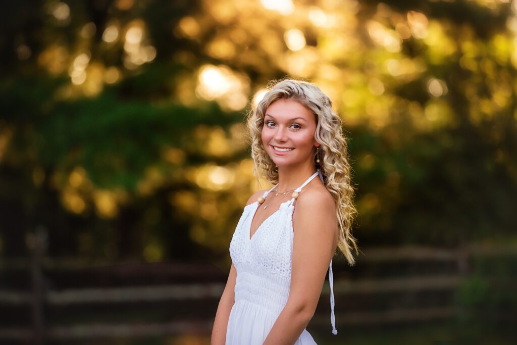 Outdoor portrait of a senior girl in a white dress, smiling with a glowing sunset background — perfect for golden hour inspiration.