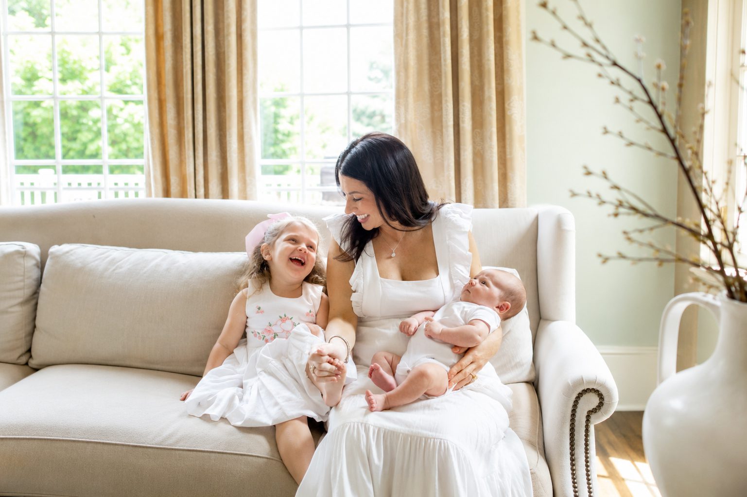 Mother cuddling newborn baby on sofa with laughing toddler during an in-home lifestyle newborn session in West Chester PA